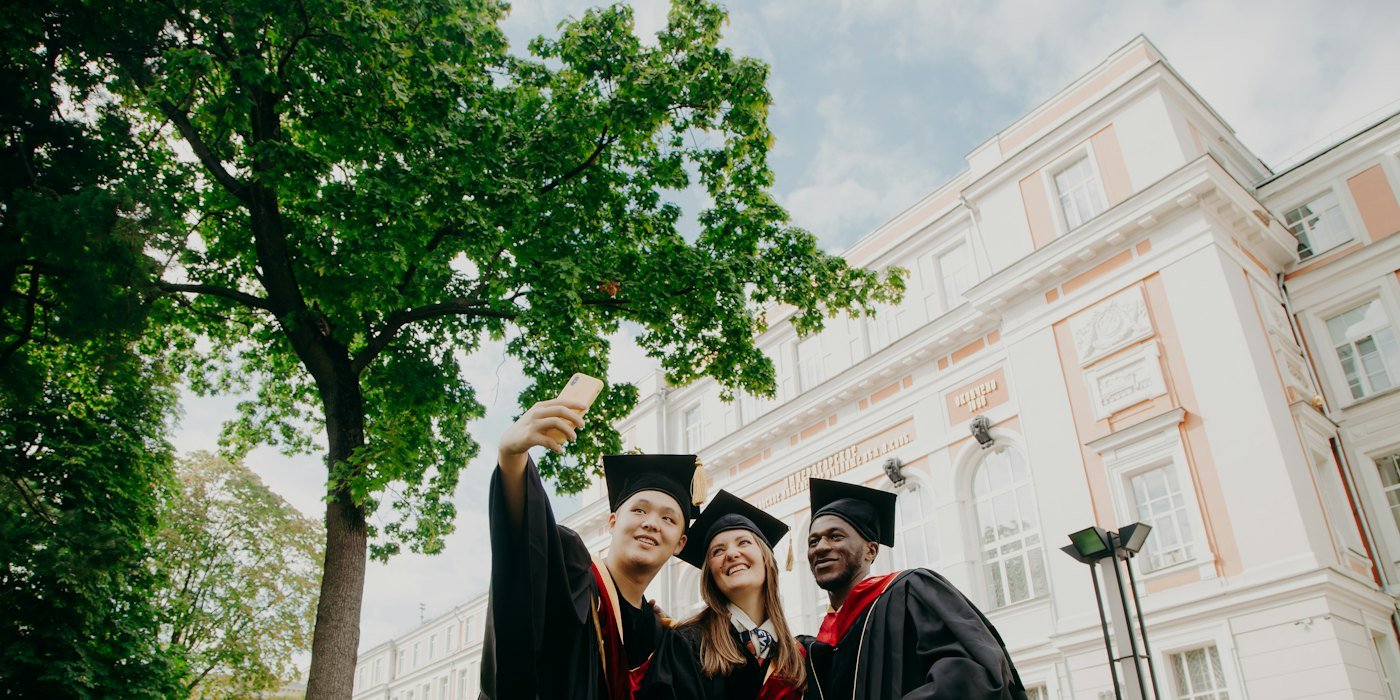 Tres graduados con toga y gorra haciéndose un selfie frente a un edificio universitario — Becas para estudiar en el extranjero